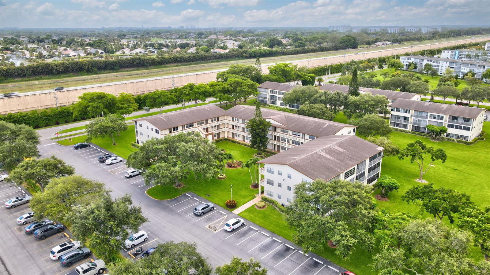 An aerial view shows a multi-story apartment complex with a parking lot and green lawns — 104 Mansfield C Drive # C, Boca Raton FL 33434, BOCA RATON, FL