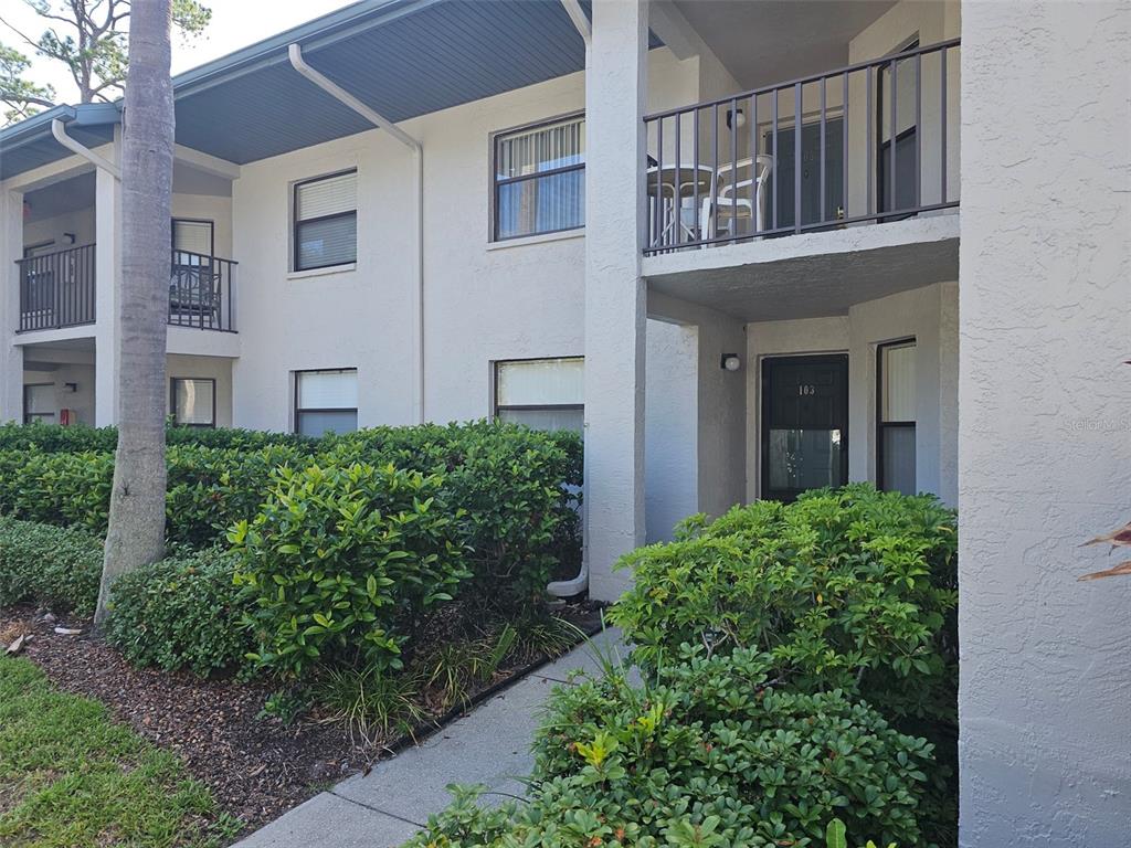 A two-story apartment building exterior features white stucco walls, dark railings, and landscaped shrubbery — 4655 W 46TH STREET COURT UNIT 103, BRADENTON, FL