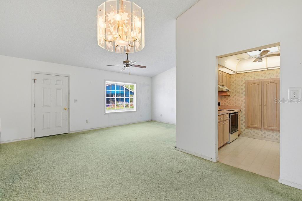 A living room with green carpeting and a chandelier opens into a kitchen with cabinets — 33 SANDRA CIRCLE, NEW SMYRNA BEACH, FL