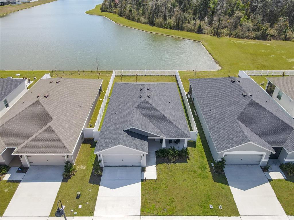 An aerial view shows three suburban houses with gray roofs and driveways facing a lake — 35598 BELLINGTON BOULEVARD, ZEPHYRHILLS, FL