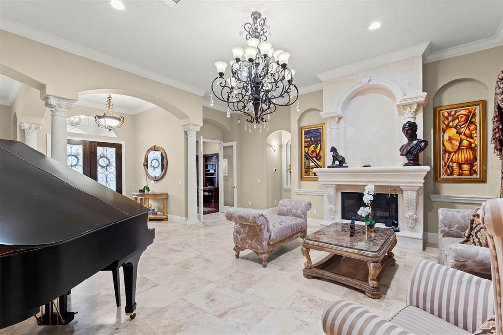 A formal living room features a grand piano, ornate chandelier, fireplace, and stone tile flooring — 7906 FOXCATCHER COURT, ODESSA, FL