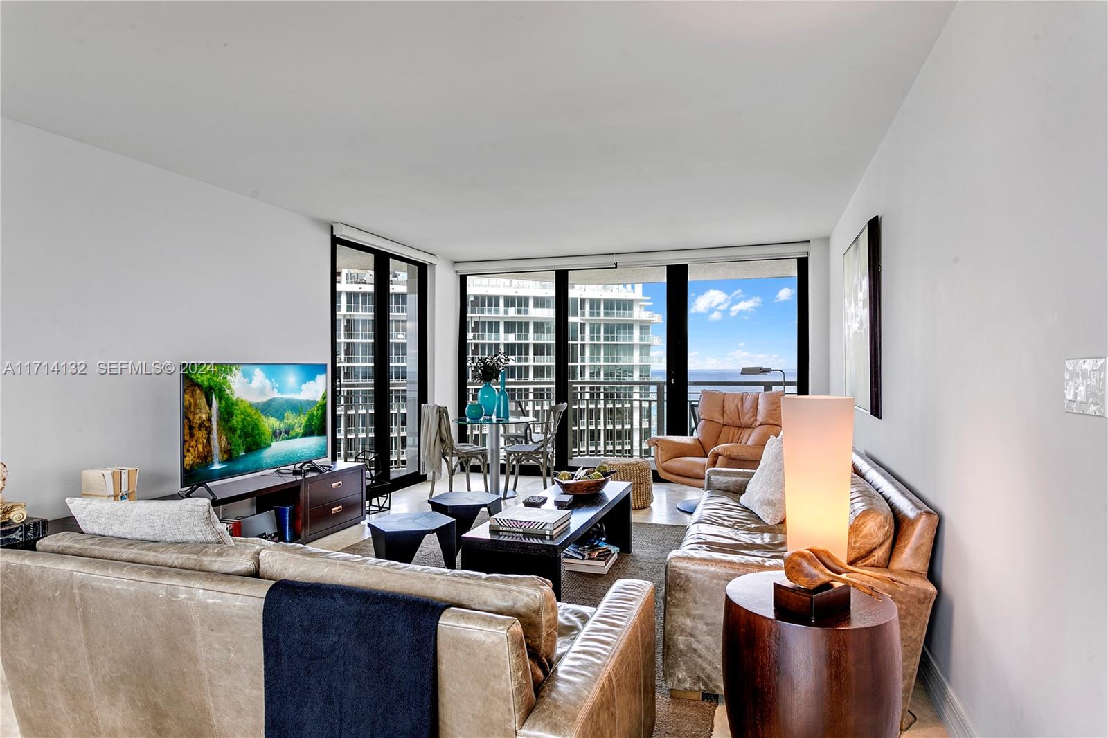 A living room features leather sofas, a television, and floor-to-ceiling windows overlooking a city skyline — 3000 N Ocean Dr # 20A, Riviera Beach FL 33404, RIVIERA BEACH, FL