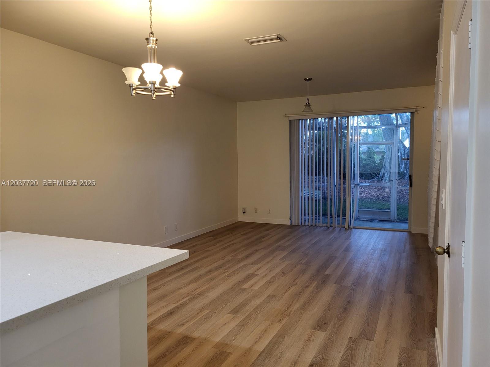 An empty room features wood-look flooring, a chandelier, and a sliding glass door to a patio — 1990 Madeira Dr, Weston FL 33327, WESTON, FL