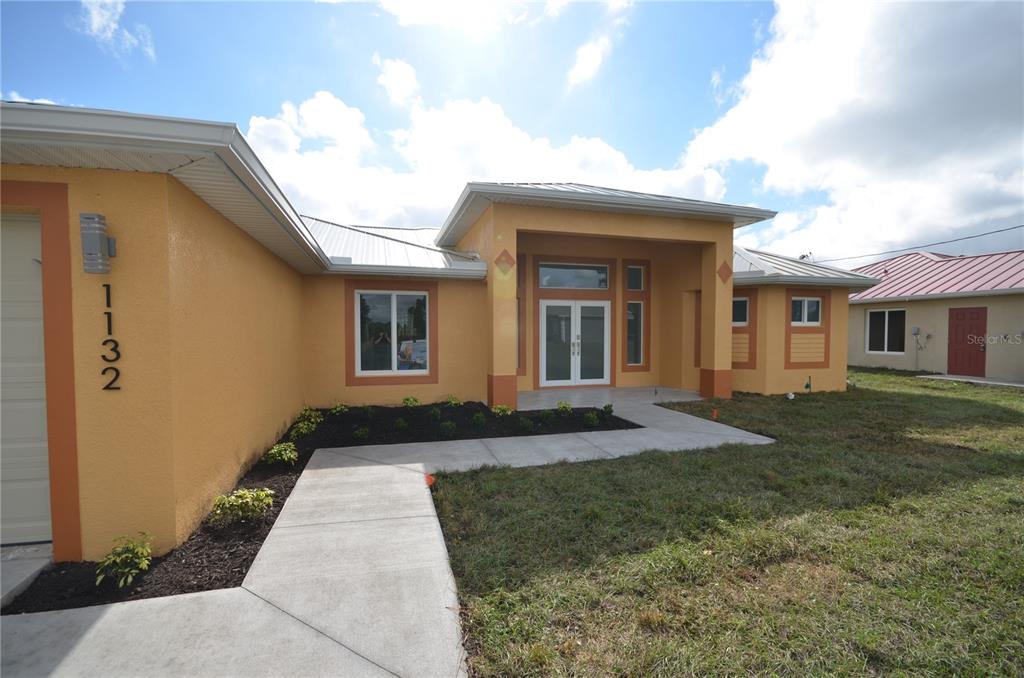 A single-story house with yellow stucco walls, a metal roof, and a concrete walkway — 1132 EDELWEISS STREET E, LEHIGH ACRES, FL