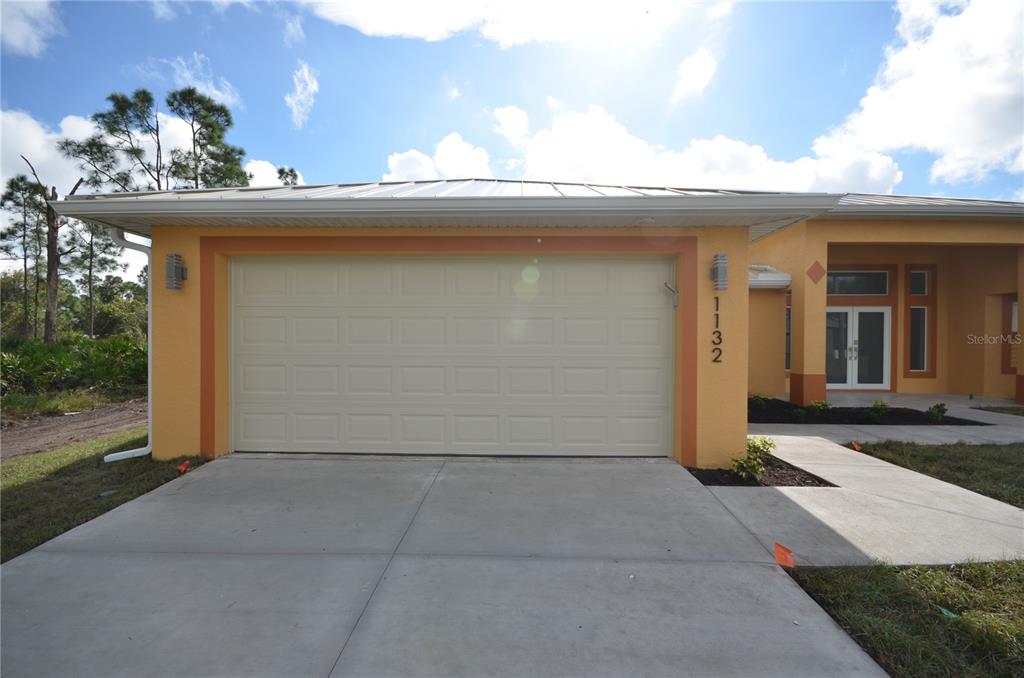 A tan house exterior features a two-car garage, a metal roof, and a concrete driveway — 1132 EDELWEISS STREET E, LEHIGH ACRES, FL