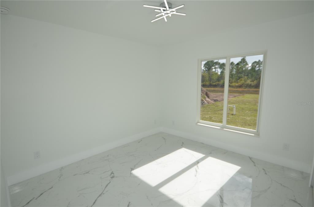 A bright, empty room features white walls, marble-patterned tile flooring, and a large window — 1132 EDELWEISS STREET E, LEHIGH ACRES, FL