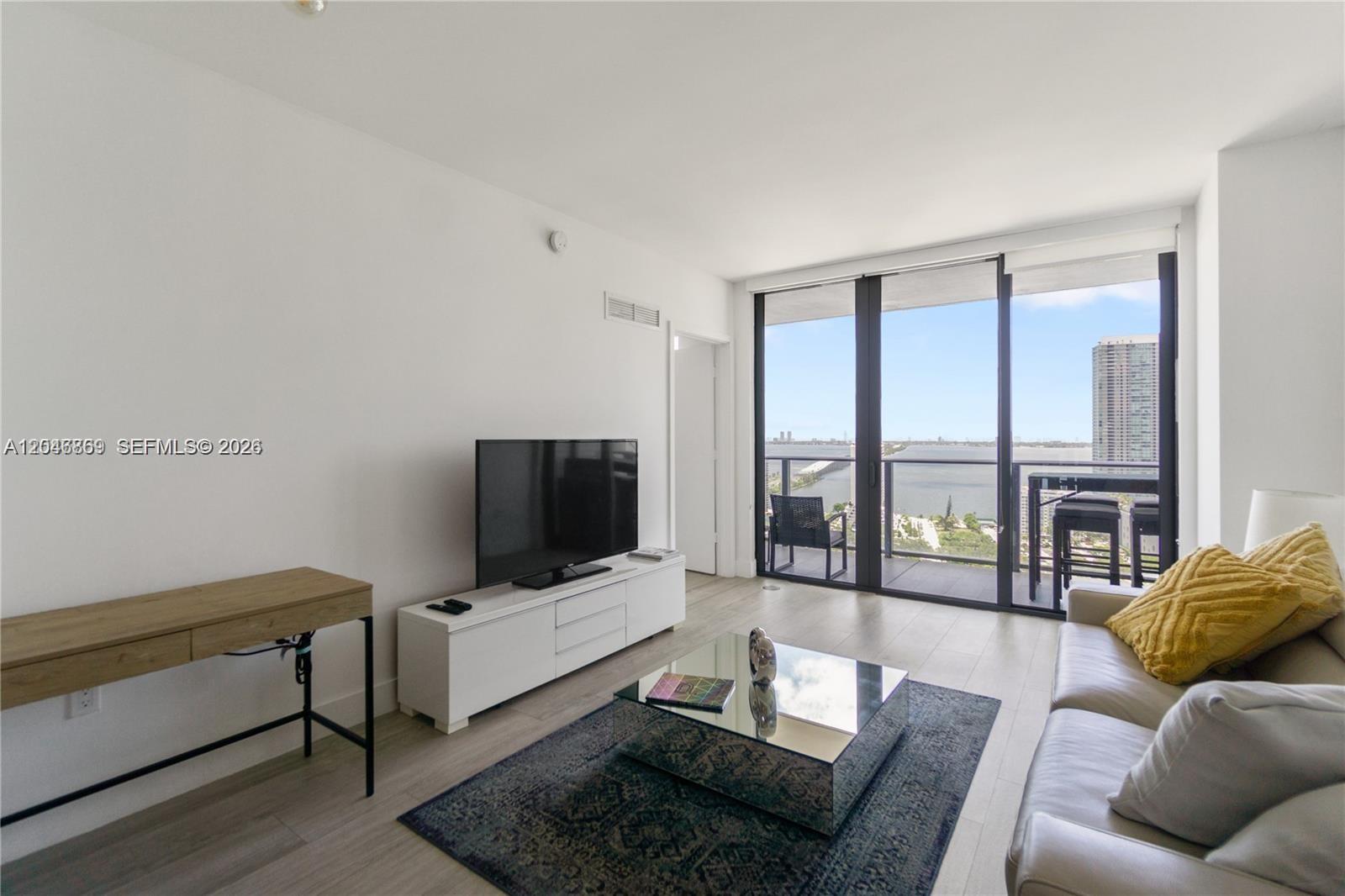 A living room features a white media console, mirrored coffee table, and balcony with water views — 121 NE 34th St # 2706, Miami FL 33137, MIAMI, FL