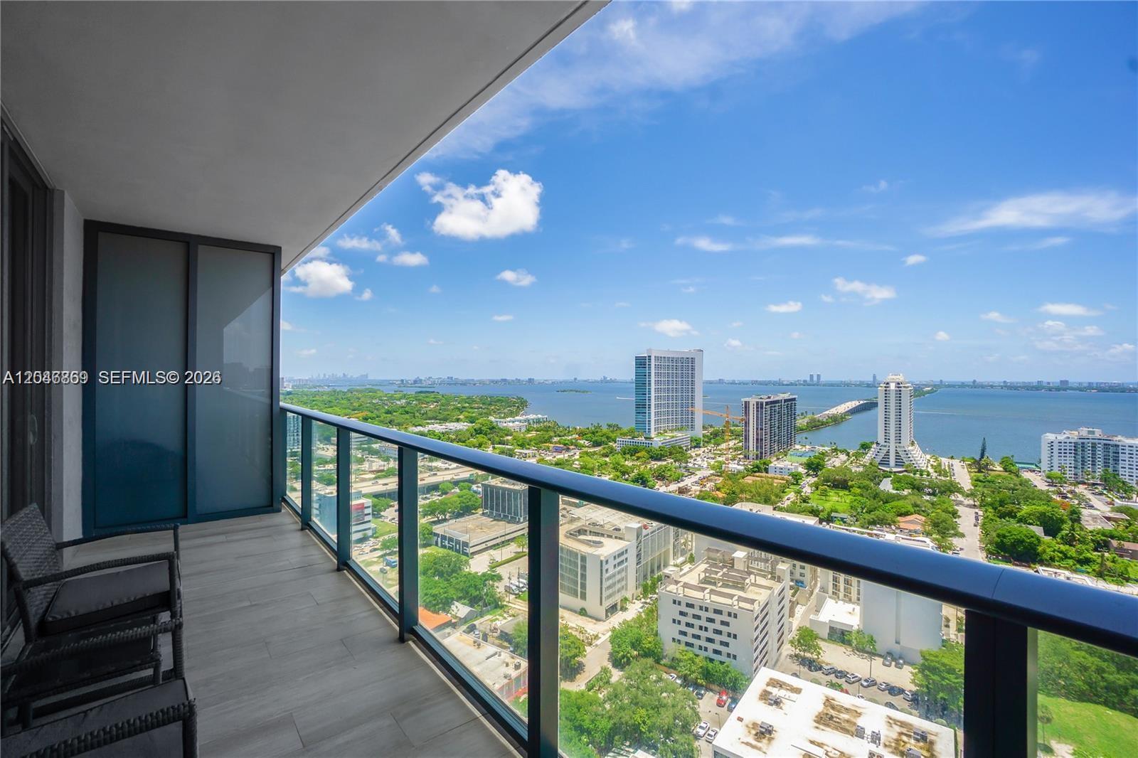 A high-rise balcony features a glass railing overlooking a city, green space, and the ocean — 121 NE 34th St # 2706, Miami FL 33137, MIAMI, FL