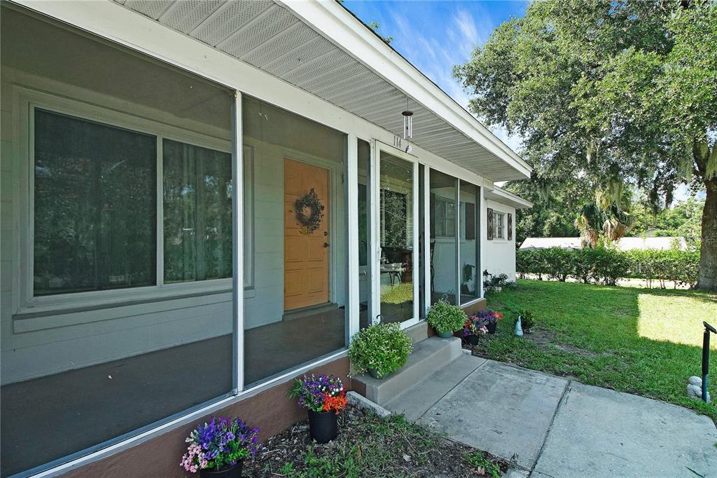 A screened-in porch with a light-colored door and potted flowers sits beside a grassy yard — 114 PARK AVENUE, FRUITLAND PARK, FL