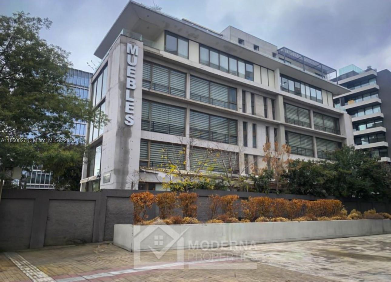 A multi-story building with a vertical sign reading MUEBLES stands behind a low concrete wall — Avda. Las Condes 10.527, 8320269, FL