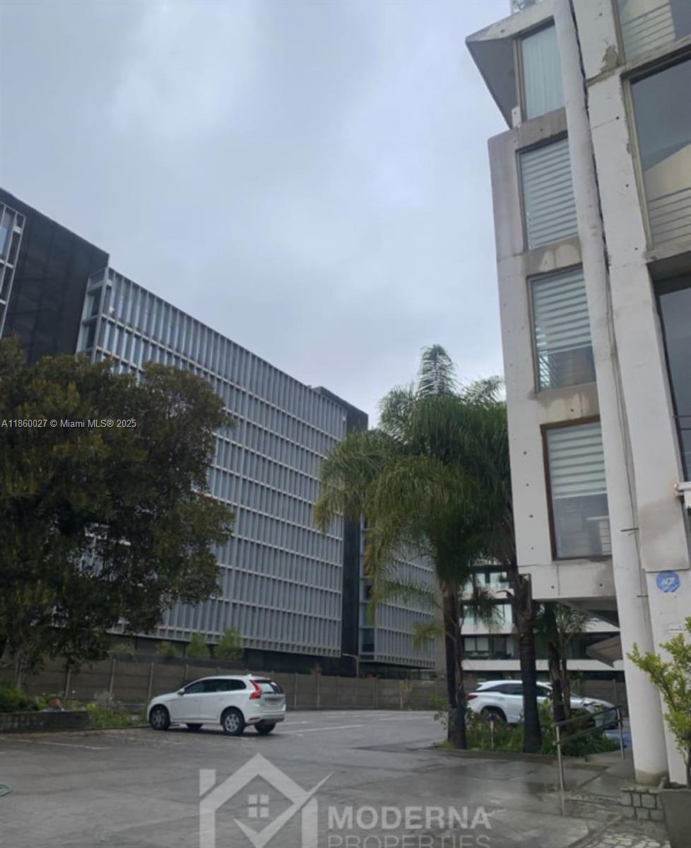 An exterior view shows a parking lot between a modern concrete building and a large office structure — Avda. Las Condes 10.527, 8320269, FL