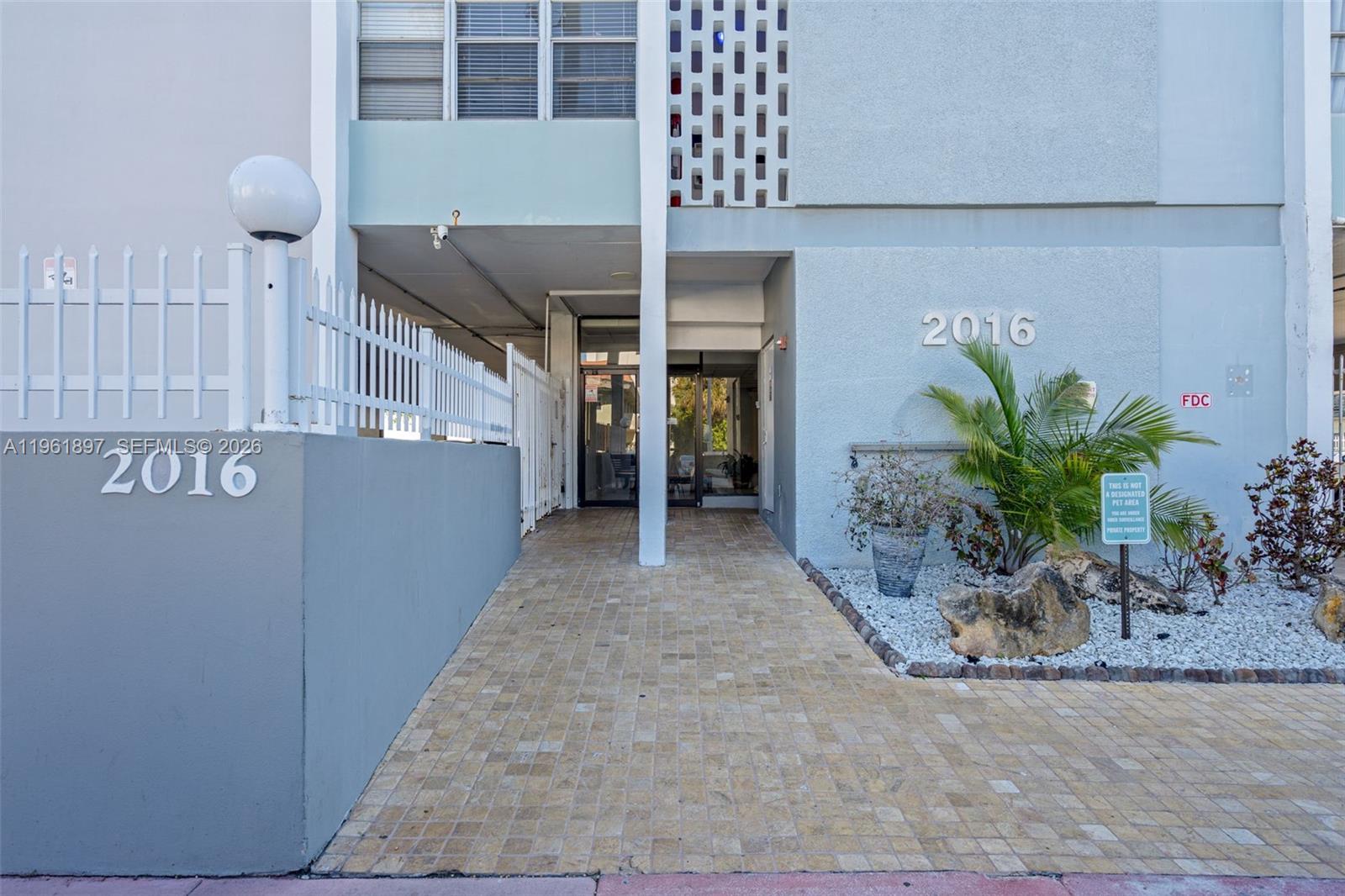 The exterior entrance of a building features a paved walkway, a white fence, and landscaping — 2016 Bay Dr # 203, Miami Beach FL 33141, MIAMI BEACH, FL