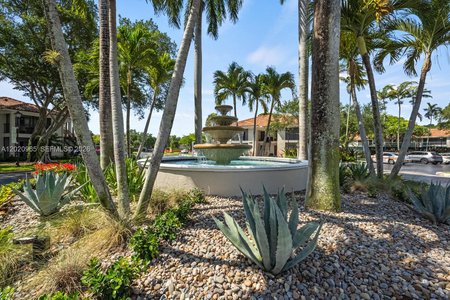 A tiered stone fountain sits in a landscaped courtyard surrounded by palm trees and buildings — 6503 N Military Trl, Boca Raton FL 33496, BOCA RATON, FL