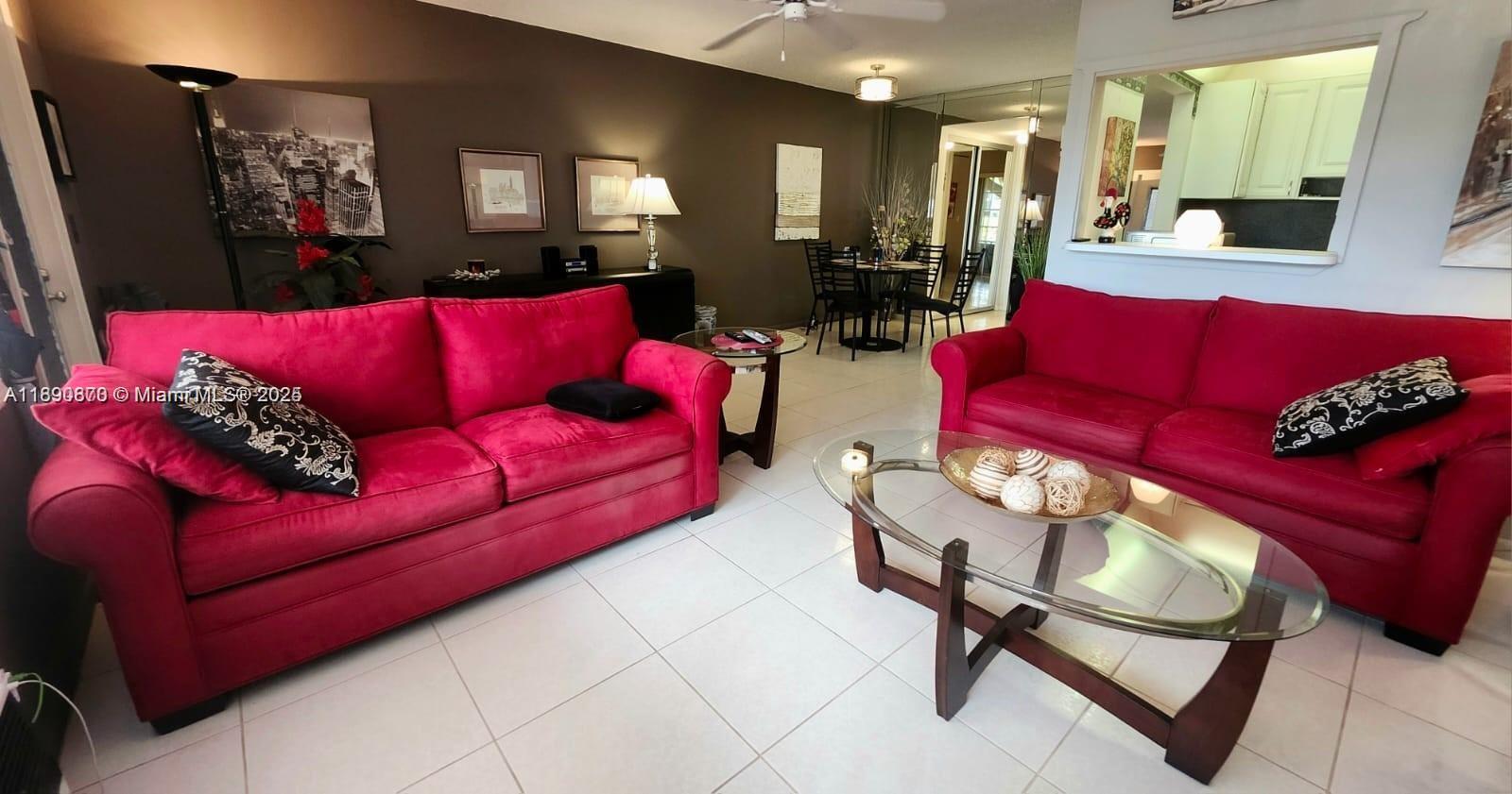 A living room features two red sofas, a glass coffee table, and tile flooring — 182 Ellesmere D # 182, Deerfield Beach FL 33442, DEERFIELD BEACH, FL