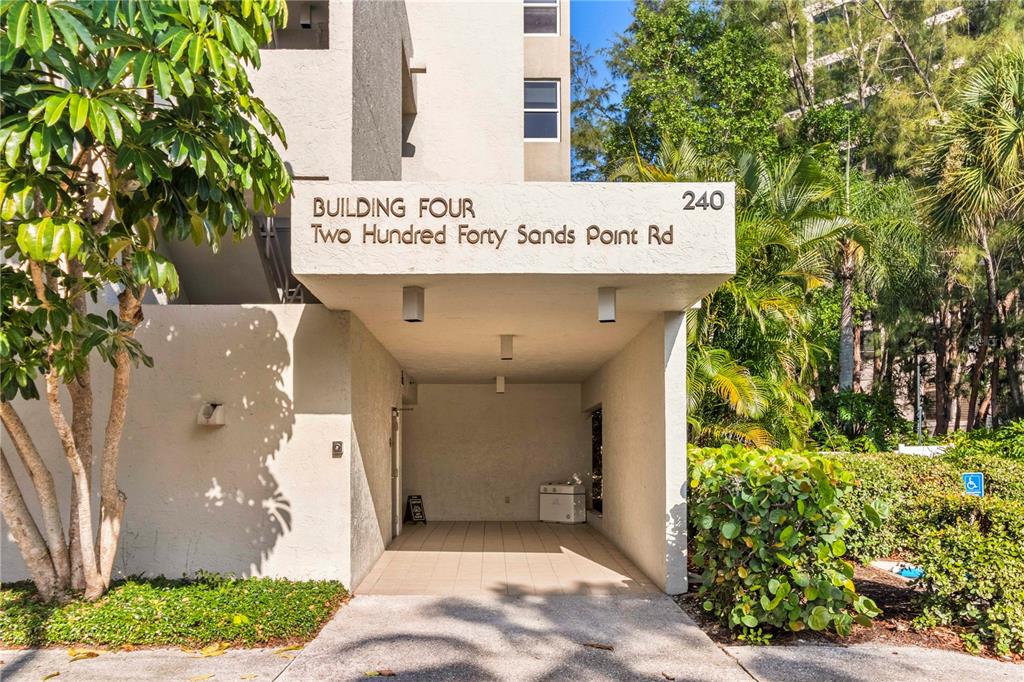 The exterior entrance of a multi-story building features a covered walkway and lush landscaping — 240 SANDS POINT ROAD UNIT 4102, LONGBOAT KEY, FL