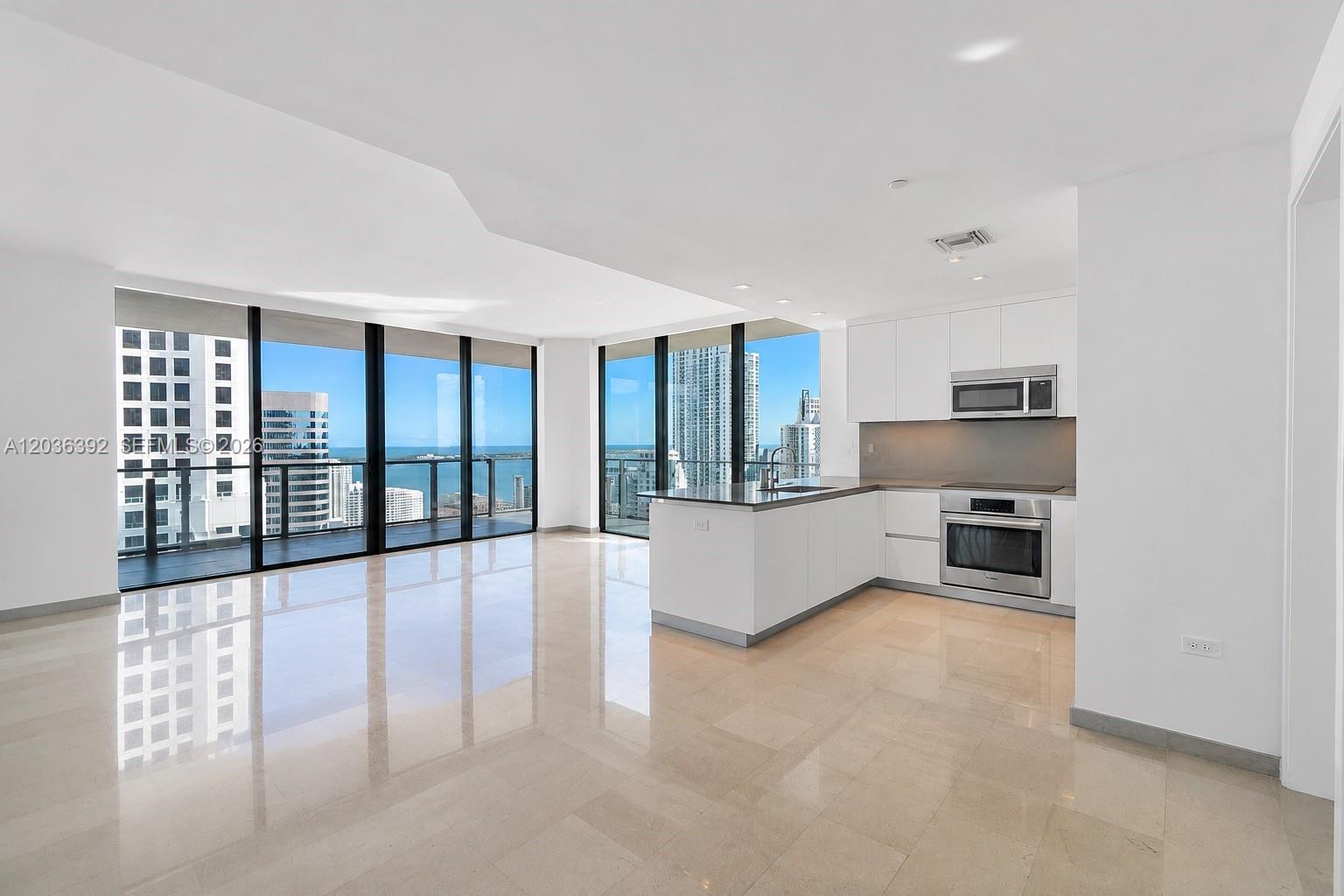 A modern kitchen with white cabinets and stainless steel appliances opens into a bright living area — 68 SE 6th St # 3301, Miami FL 33131, MIAMI, FL