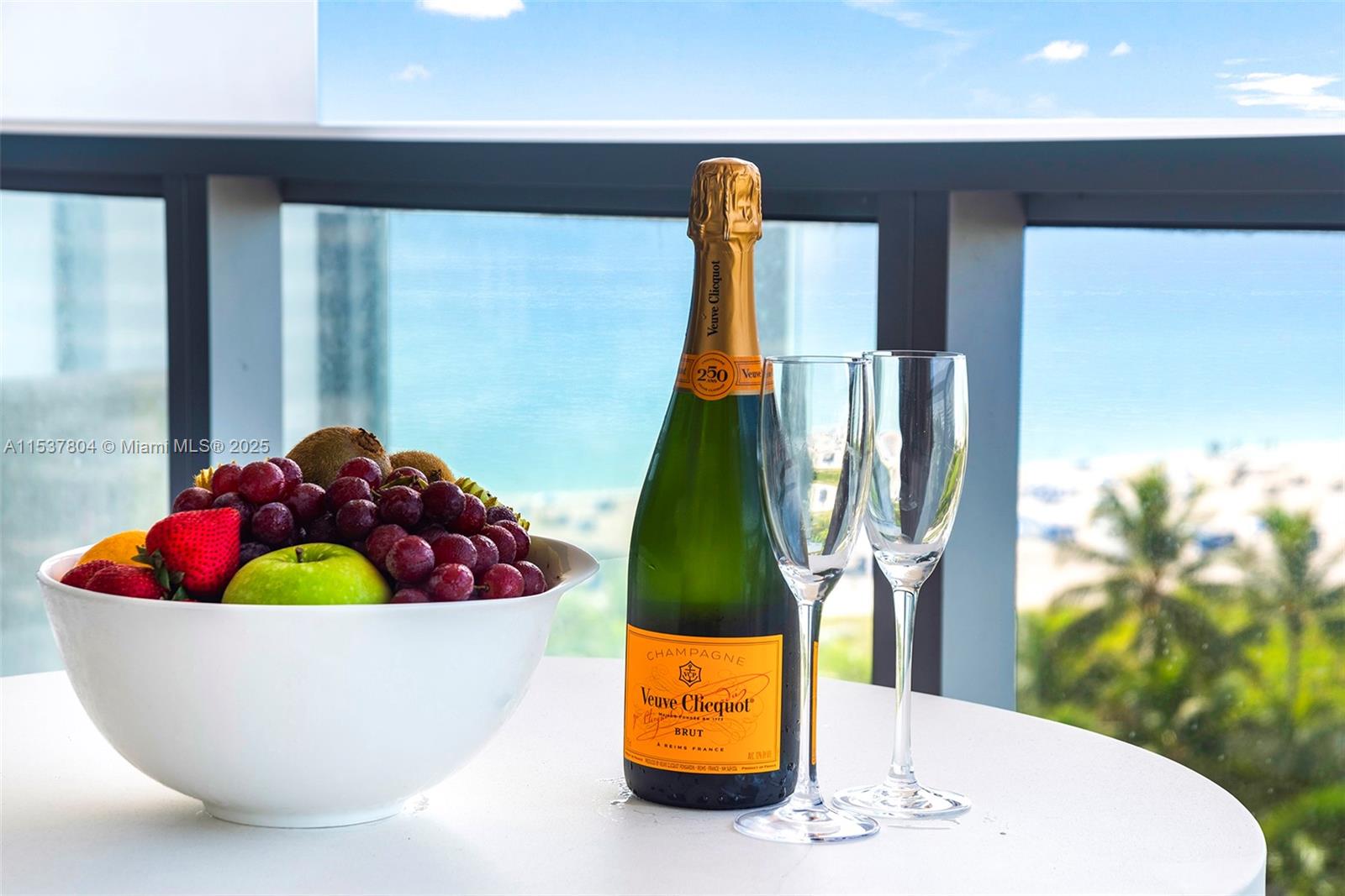 A bowl of fruit and a bottle of champagne sit on a table overlooking the ocean — 2201 Collins Ave # 814, Miami Beach FL 33139, MIAMI BEACH, FL