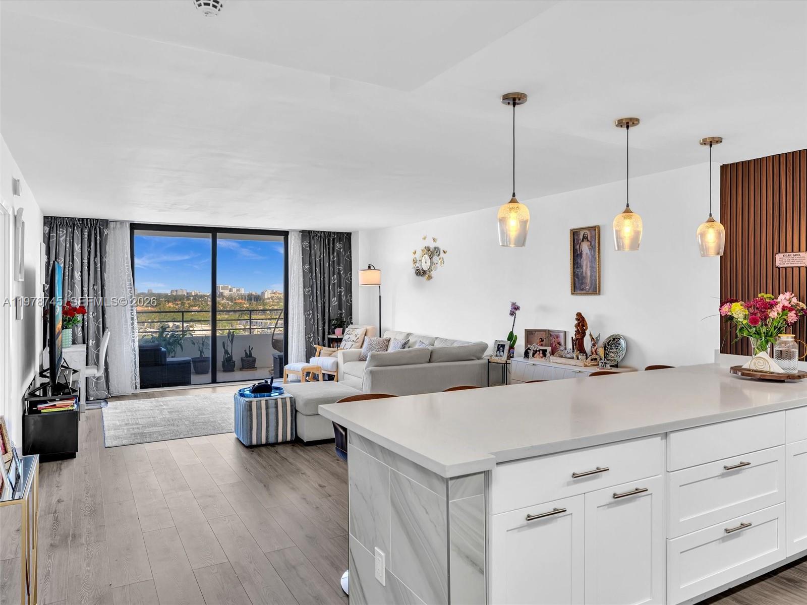 A modern open-concept living room and kitchen with white cabinetry, wood flooring, and balcony access — 2500 Parkview Dr # 1108, Hallandale Beach FL 33009, HALLANDALE BEACH, FL