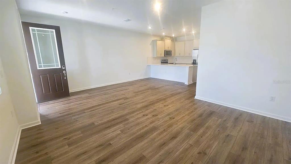 An open-concept living area features wood-look flooring, a dark front door, and a kitchen — 7774 CLOVE HITCH WAY, ORLANDO, FL