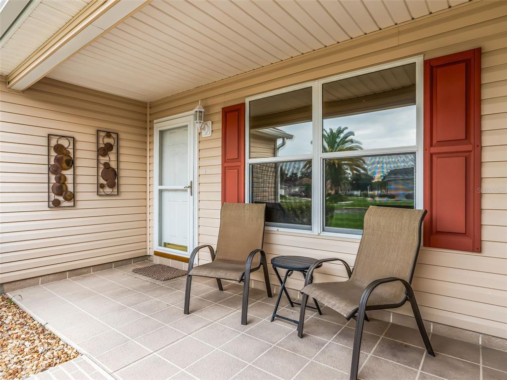A covered porch features tan siding, a white storm door, two chairs, and a small table — 1502 AQUINO LANE, THE VILLAGES, FL