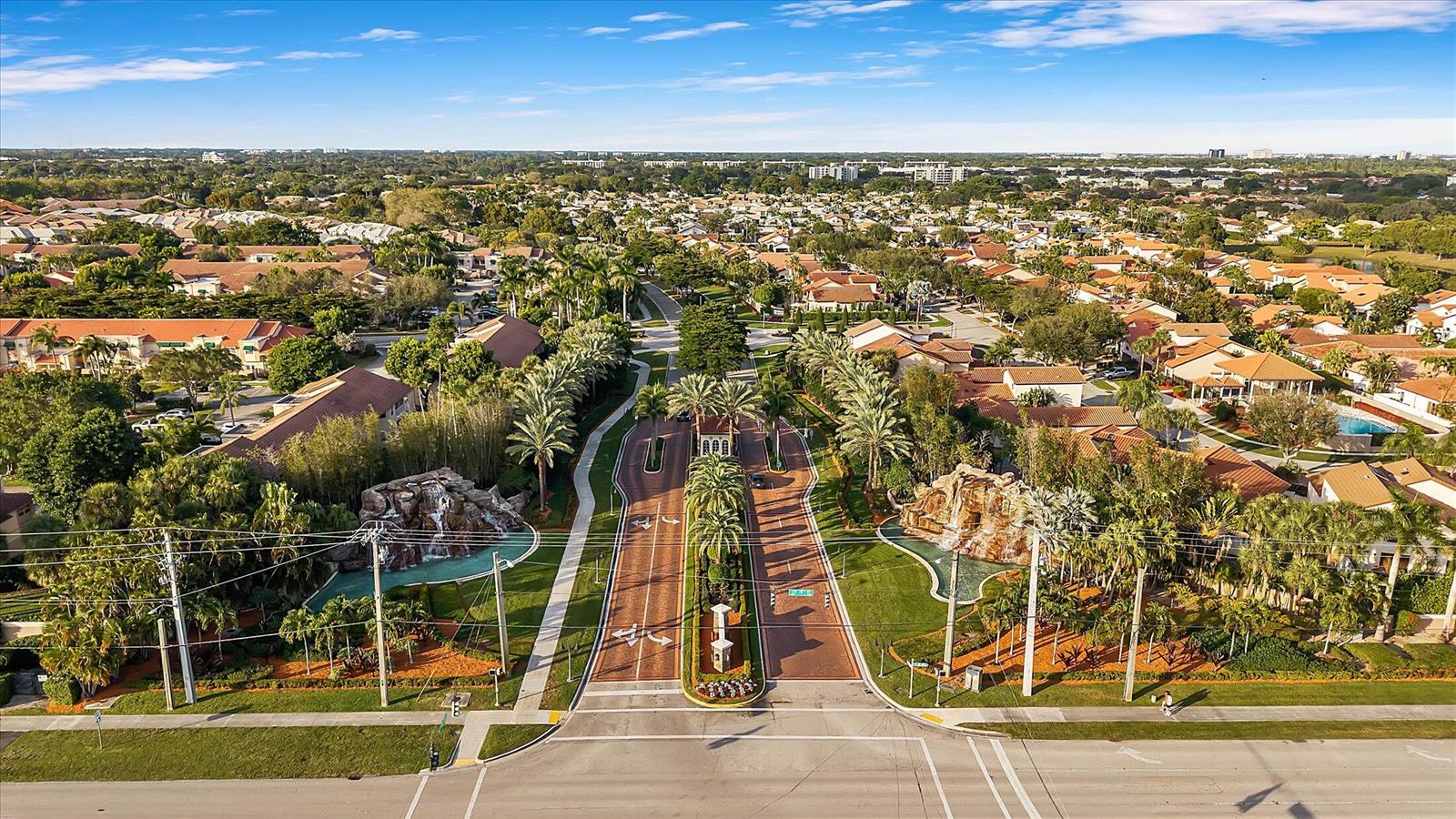 An aerial view of a gated community entrance featuring two stone waterfalls and palm trees — 6757 Via Regina # 3, Boca Raton FL 33433, BOCA RATON, FL