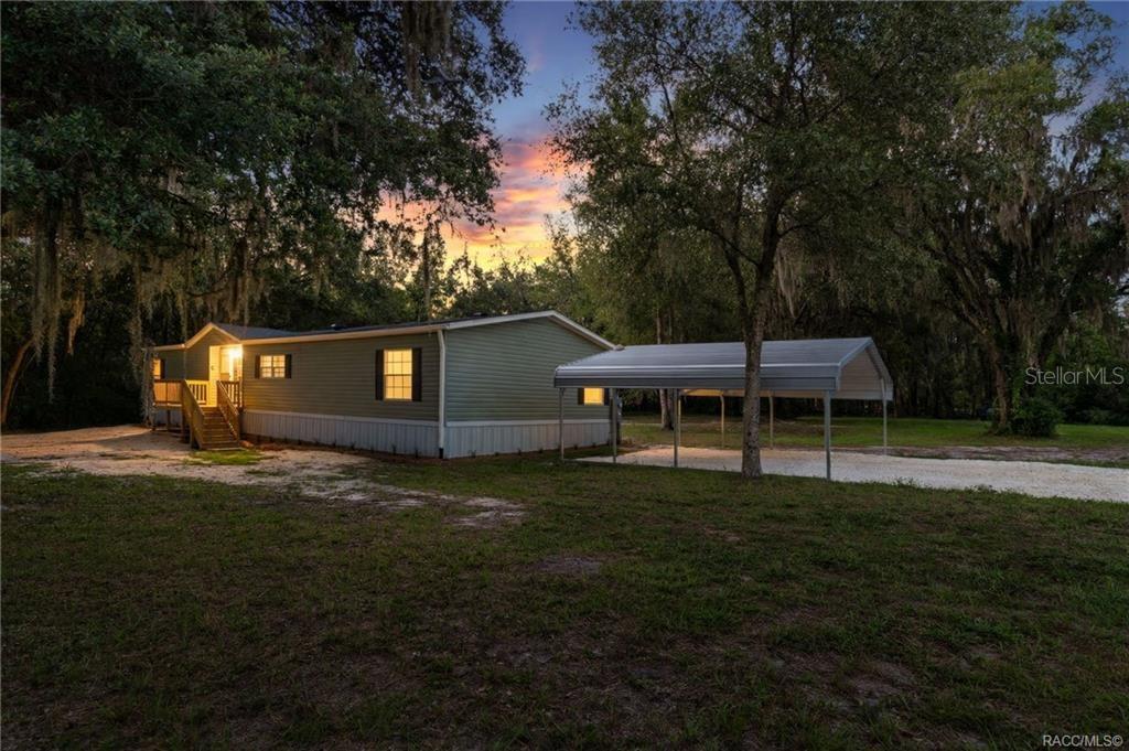 A light green manufactured home with a metal carport sits on a grassy, wooded lot — 11437 N YOUNG DRIVE, DUNNELLON, FL