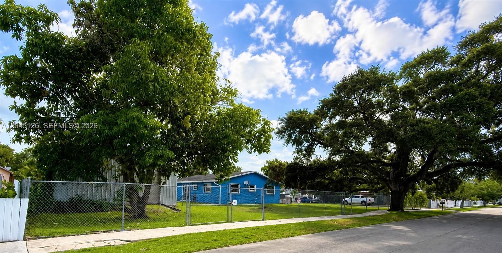 A blue house sits behind a chain-link fence surrounded by large trees and grass — 6145 Hayes St, Hollywood FL 33024, HOLLYWOOD, FL