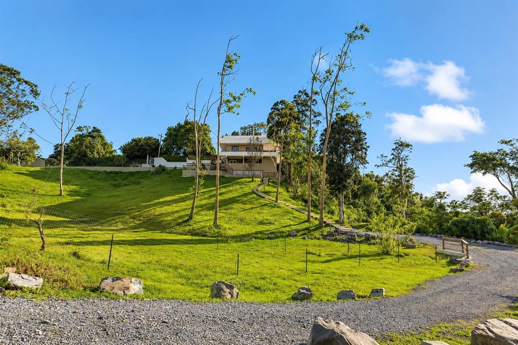 A house sits atop a grassy hill with a gravel driveway in the foreground — RD15 KM20.6 BO. QUEBRADA ARRIBA, CAYEY, PR