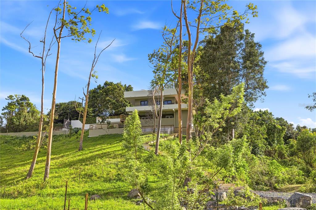 A modern multi-story house sits on a grassy hillside surrounded by trees and vegetation — RD15 KM20.6 BO. QUEBRADA ARRIBA, CAYEY, PR