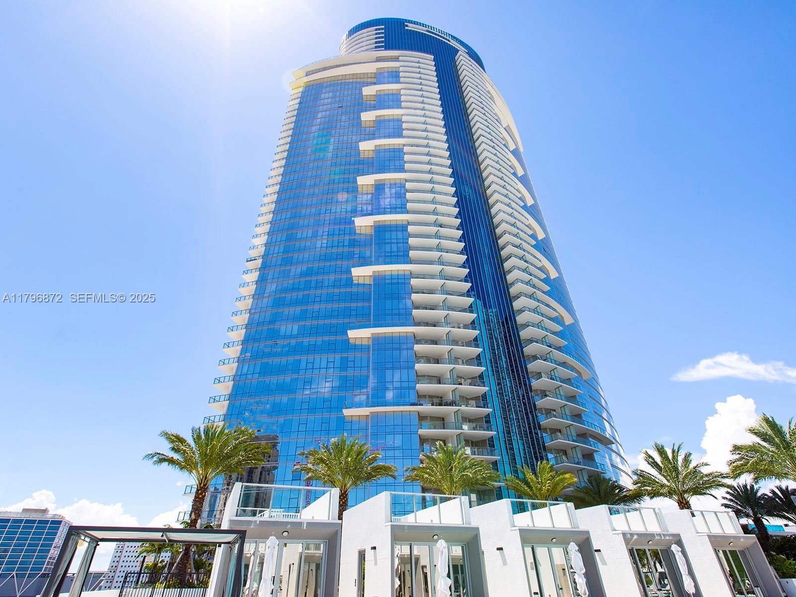 A low-angle view of a modern blue glass skyscraper with curved balconies and palm trees — 851 NE 1st Ave, Miami FL 33132, MIAMI, FL