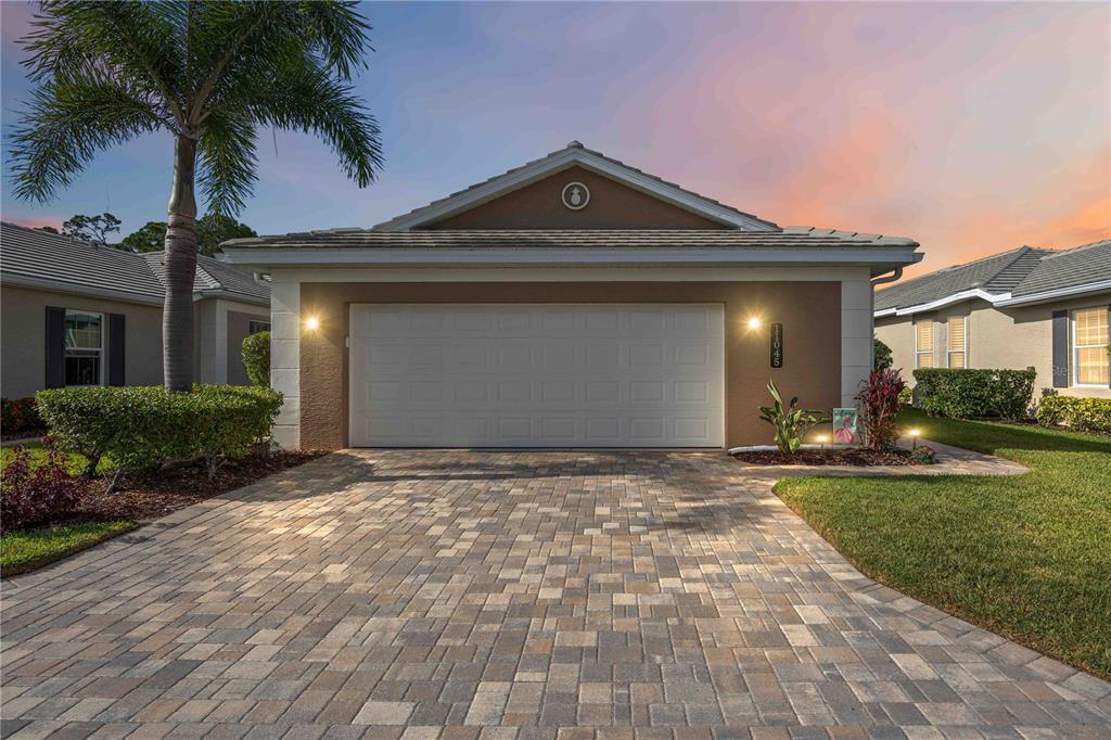 A single-story house with a two-car garage, paver driveway, and a palm tree at sunset — 11045 BARNSLEY DRIVE, VENICE, FL
