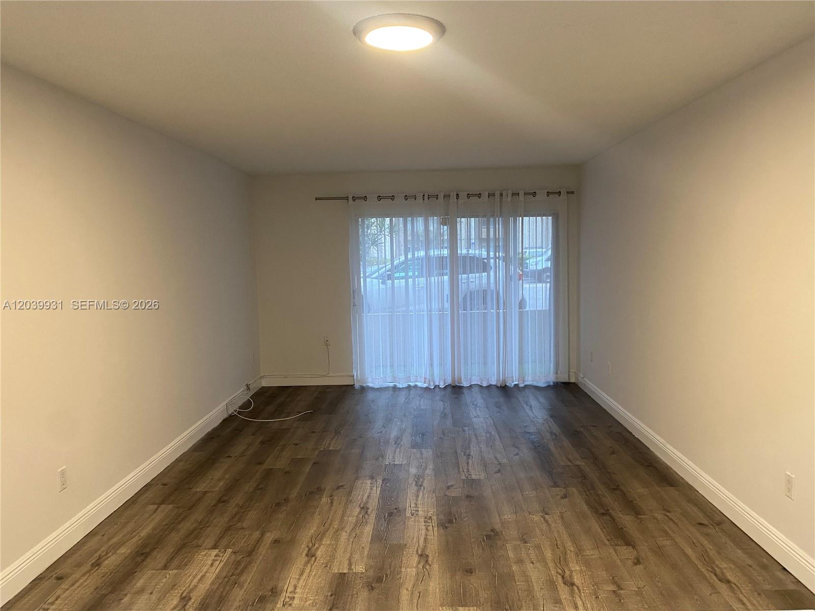 An empty room features wood-look laminate flooring, white walls, and a sliding glass door — 7755 SW 86th St # 107, Miami FL 33143, MIAMI, FL