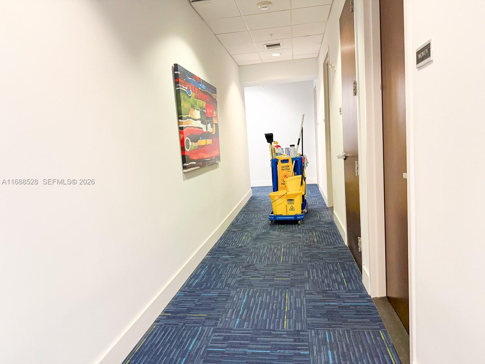 A hallway with blue patterned carpet, white walls, abstract art, and a janitorial cart — 2020 Ponce De Leon Blvd, Coral Gables FL 33134, CORAL GABLES, FL