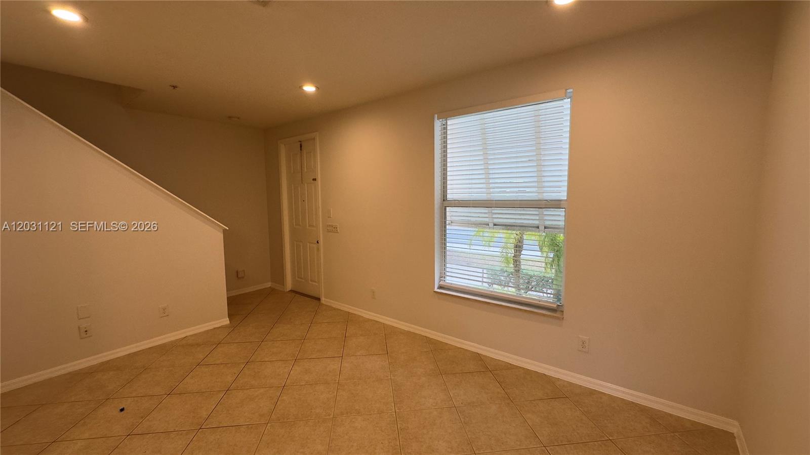 An empty room features beige tile flooring, a staircase, a closed door, and a window — 1587 Frederick Small Rd, Jupiter FL 33458, JUPITER, FL