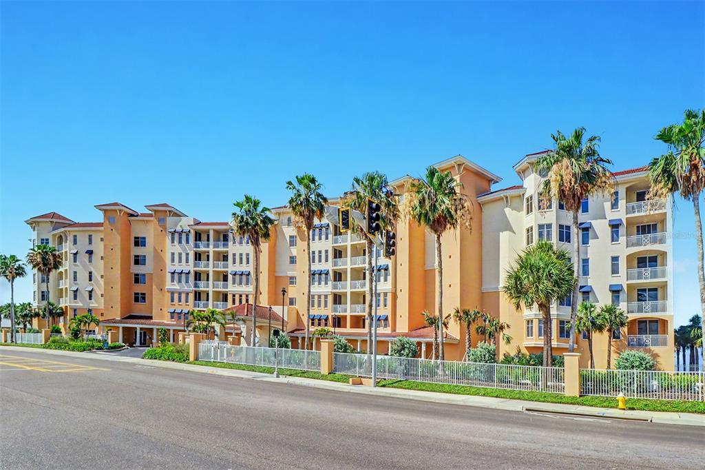 A multi-story tan apartment building with balconies and palm trees sits beside a paved road — 15208 GULF BOULEVARD UNIT 601, MADEIRA BEACH, FL