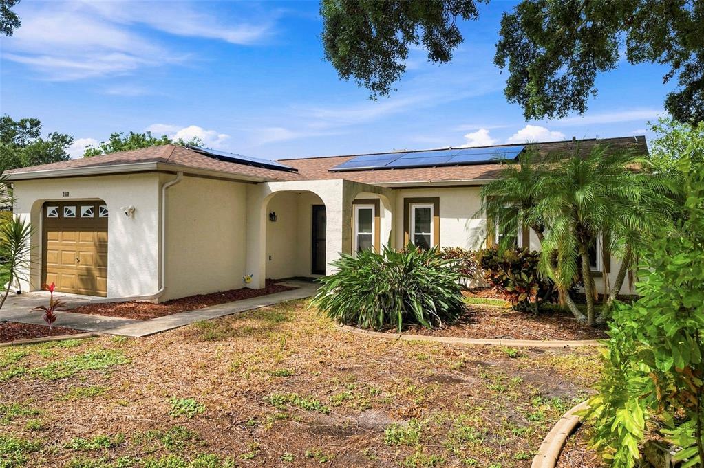 A single-story house with a tan stucco exterior, a brown garage door, and rooftop solar panels — 2460 BEACON GROVES BOULEVARD, PALM HARBOR, FL