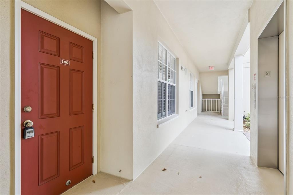 A red apartment door with a lockbox sits on a covered outdoor walkway near an elevator — 855 BLUE SAGE STREET UNIT 102, CELEBRATION, FL