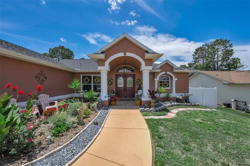 A tan house exterior features a covered entryway, red canna lilies, and a stone chimney — 11210 AUBURNDALE STREET, SPRING HILL, FL