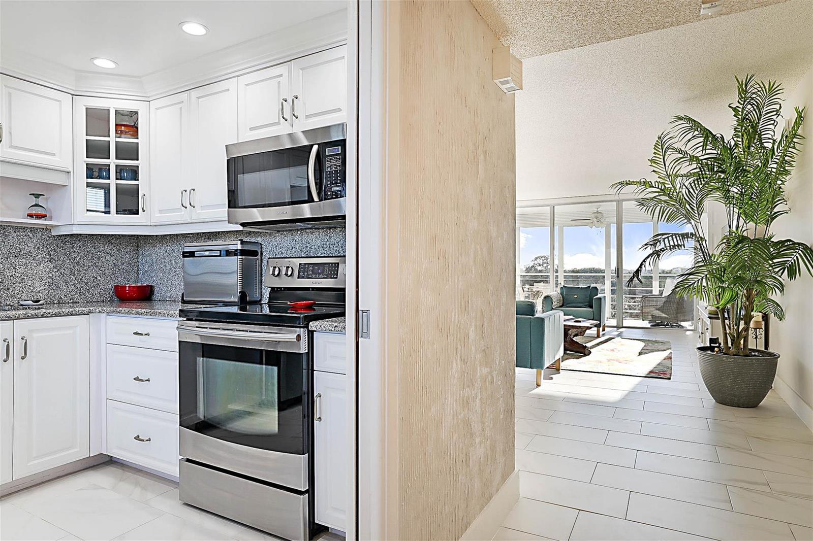 A white kitchen with stainless steel appliances opens into a living area with a plant — 575 Oaks Lane # 703, Pompano Beach FL 33069, POMPANO BEACH, FL