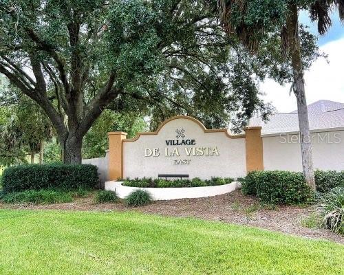 A beige monument sign for Village De La Vista East stands in a grassy yard — 1709 SONORA STREET, THE VILLAGES, FL