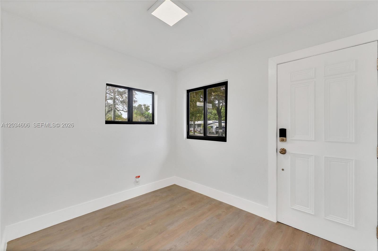 A small, empty room features white walls, light wood-look flooring, two black-framed windows, and a white door — 2428 NW 28th St, Miami FL 33142, MIAMI, FL