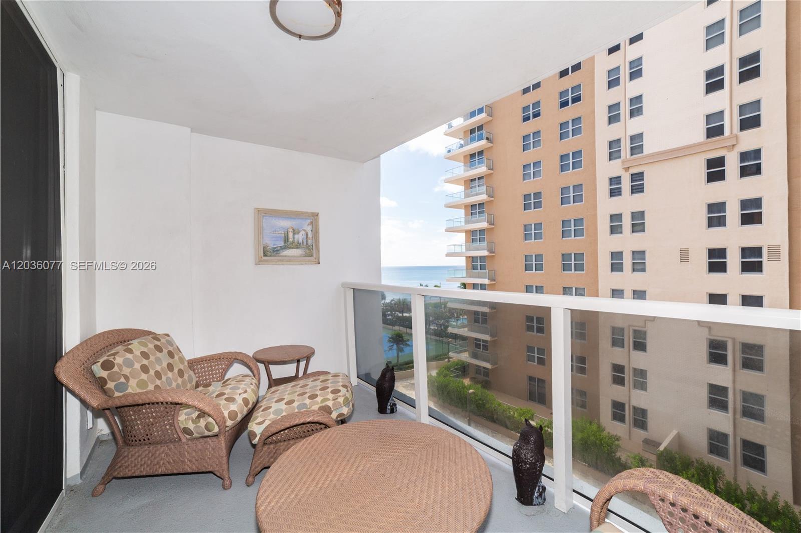 A balcony features wicker furniture, a small painting, and a view of a neighboring building — 1904 S Ocean Dr # 604, Hallandale Beach FL 33009, HALLANDALE BEACH, FL