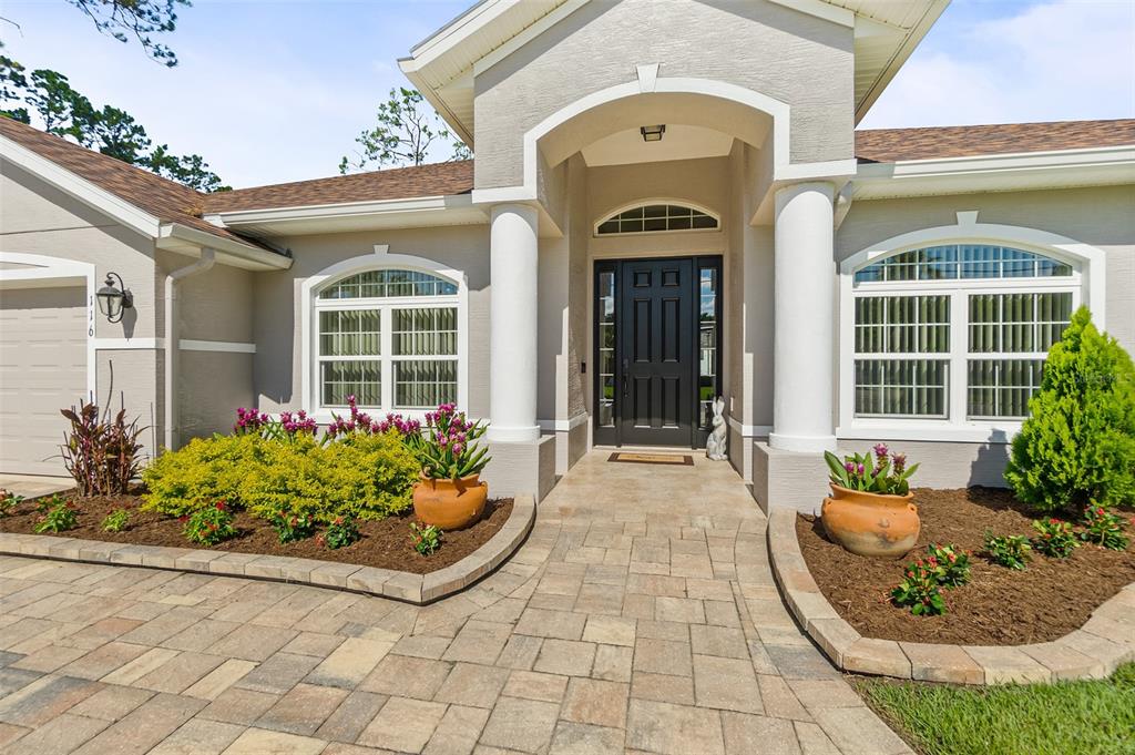 A beige house exterior features a paved walkway, a black front door, and landscaped garden beds — 116 ERIC DRIVE, PALM COAST, FL