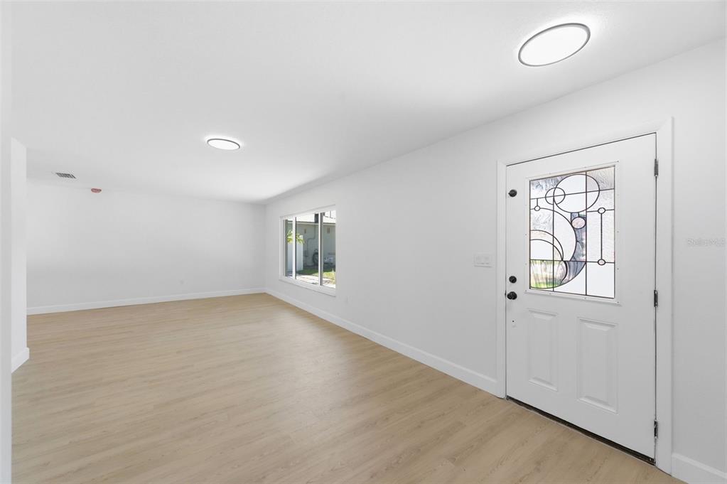 A bright, empty living room features light wood-look flooring, white walls, and a decorative front door — 11180 113TH AVENUE, LARGO, FL