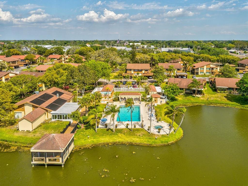 An aerial view shows a community swimming pool and clubhouse beside a lake and buildings — 3843 LIGHTHOUSE WAY, NEW PORT RICHEY, FL