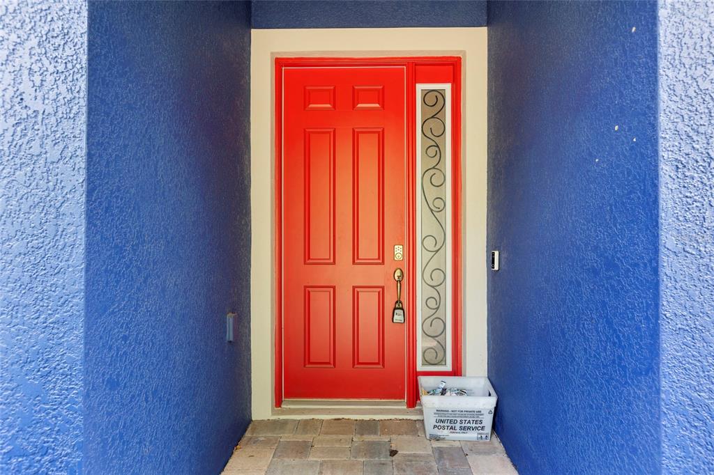A bright red front door with a decorative glass sidelight sits between blue textured walls — 7013 ARTESIAN COURT, APOLLO BEACH, FL
