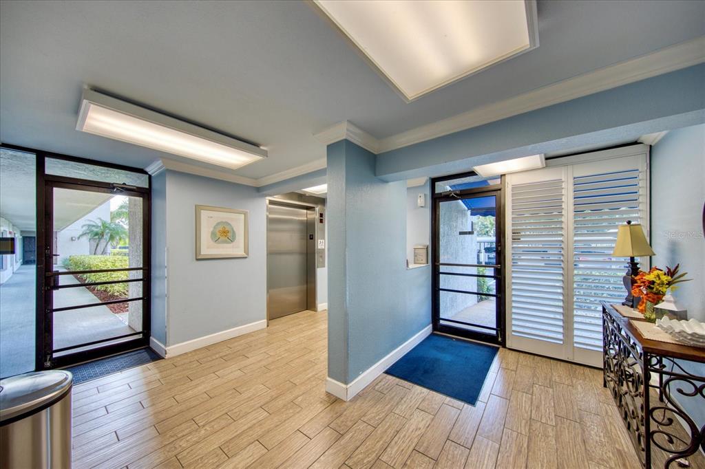 A building lobby features light blue walls, wood-look tile flooring, an elevator, and glass doors — 1324 PASADENA AVENUE S UNIT 204, SOUTH PASADENA, FL