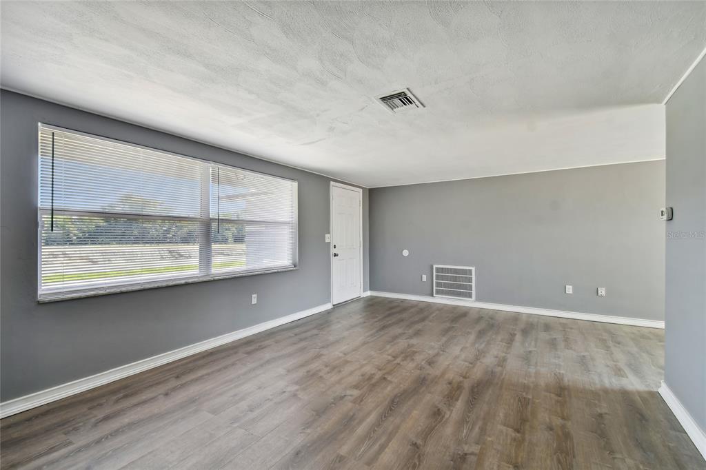 An empty room with gray walls, wood-look flooring, a large window with blinds, and a door — 7721 ARBORDALE DRIVE, PORT RICHEY, FL