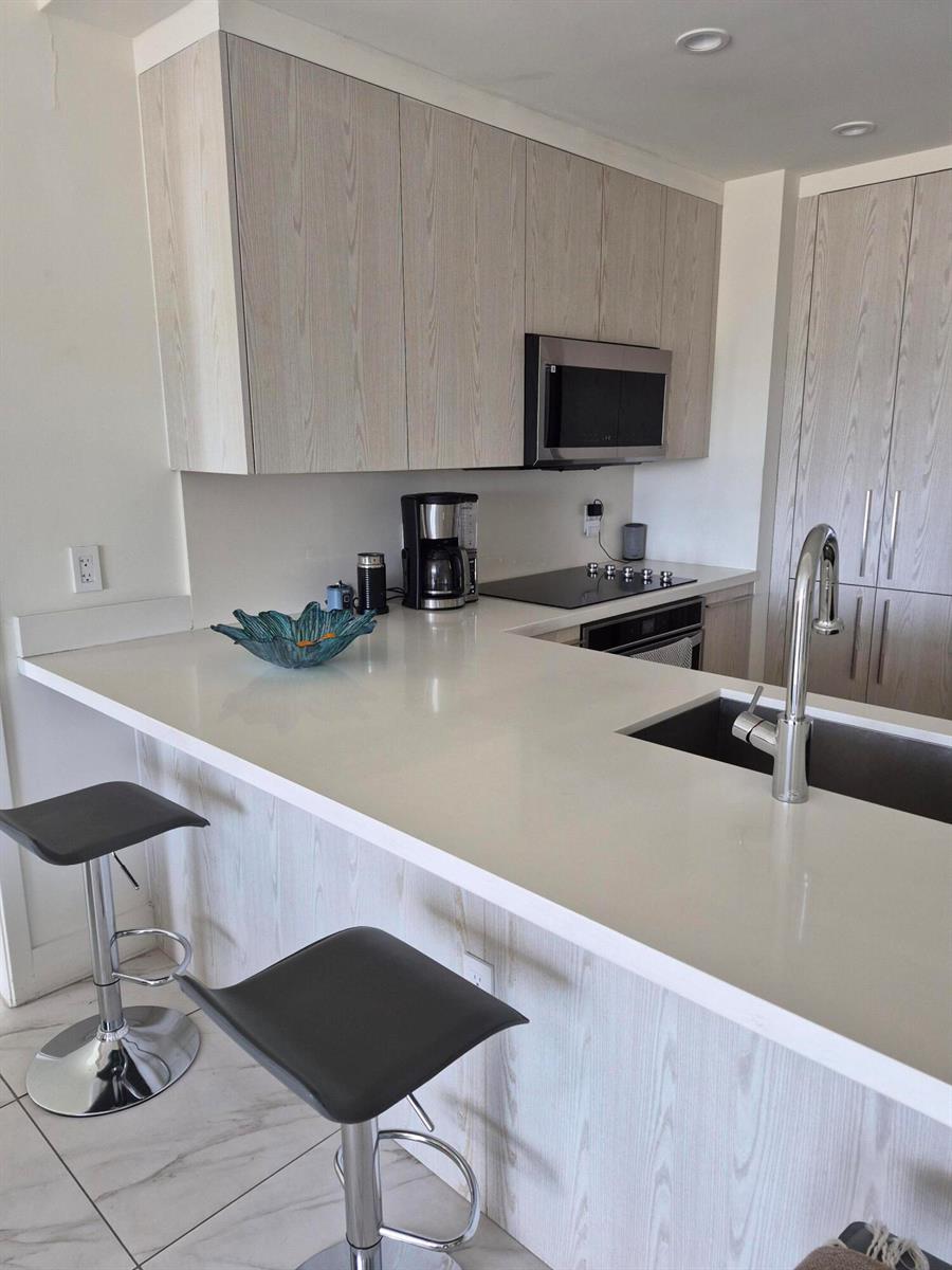 A modern kitchen features light wood cabinets, white countertops, a breakfast bar, and two stools — 2000 Metropica Way # 507, Sunrise FL 33323, SUNRISE, FL