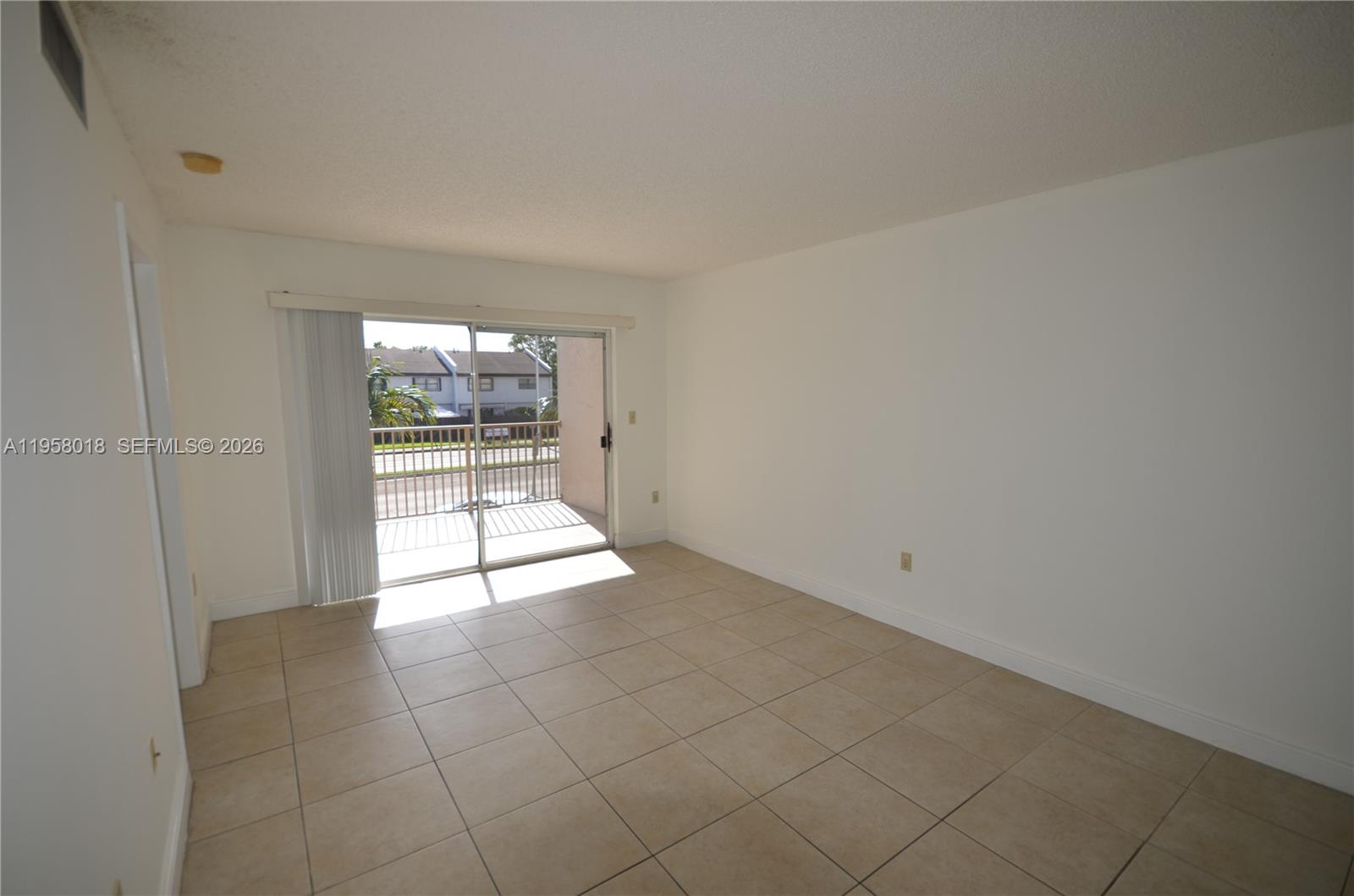 A bright, empty room features beige tile flooring, white walls, and a sliding glass balcony door — 15201 SW 80th St # 207, Miami FL 33193, MIAMI, FL